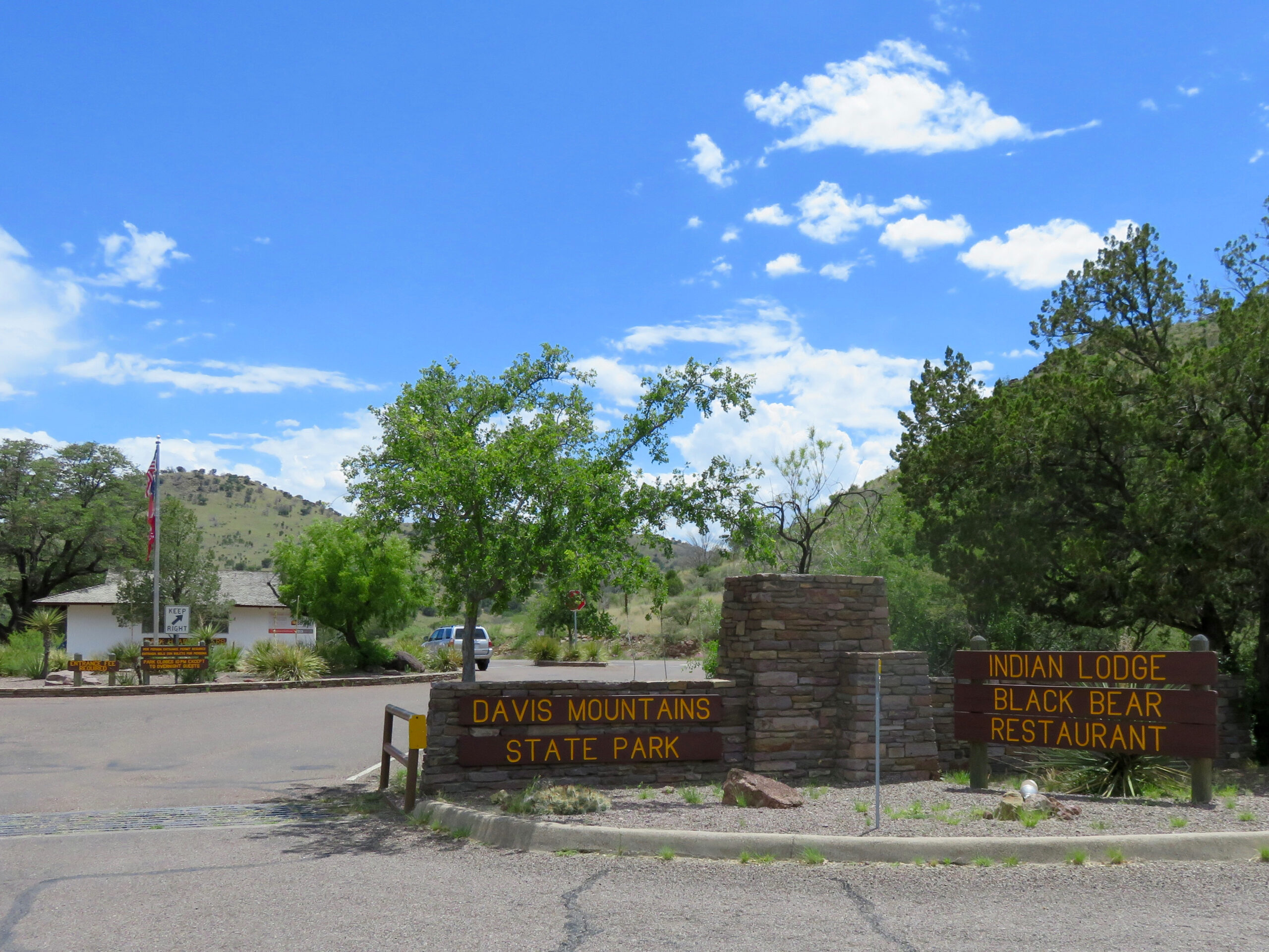 Entrance to Davis Mountains State Park with signage for the Indian Lodge Black Bear Restaurant, surrounded by trees and hills under a blue sky with clouds.