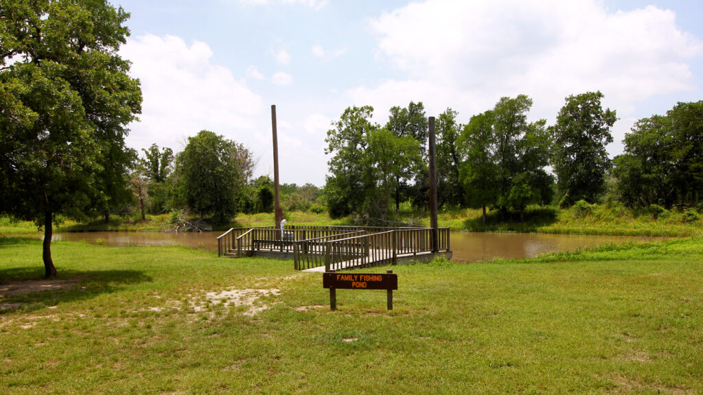 A wooden dock extends into a small pond surrounded by grass and trees. A sign labeled "Family Fishing Pond" is positioned in the foreground. The sky is partly cloudy.