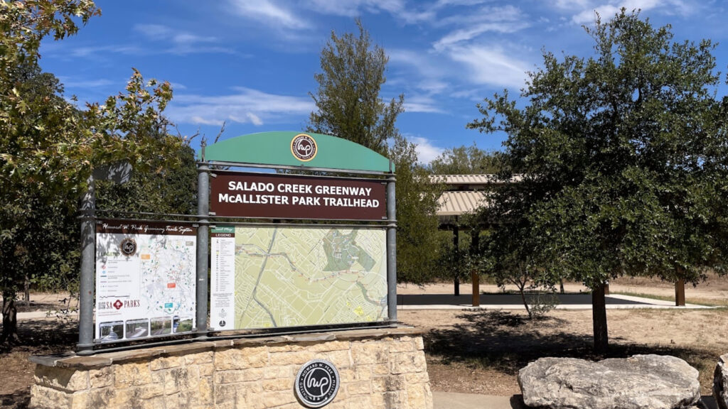A trailhead sign at McAllister Park for the Salado Creek Greenway is displayed, with maps and information boards, surrounded by trees under a partly cloudy sky.