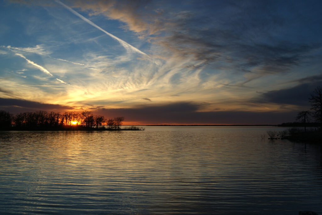 Sun setting over a calm body of water, with silhouetted trees on the horizon and wispy clouds streaking across the sky.