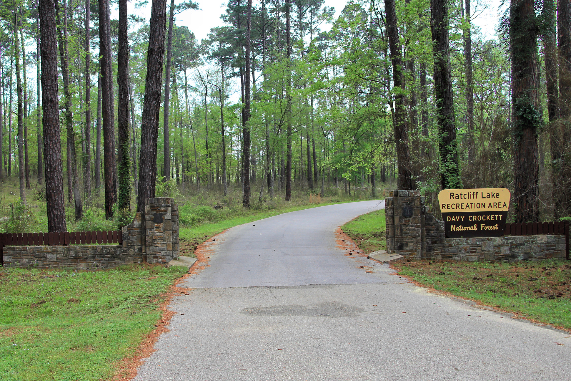 Paved road flanked by stone pillars leading through a forest with a sign on the right reading "Ratcliff Lake Recreation Area, Davy Crockett National Forest.
