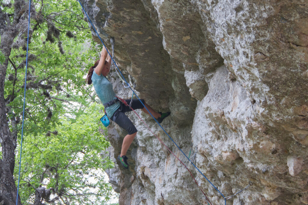 A person wearing climbing gear is scaling a rock formation at Reimers Ranch, surrounded by trees and using ropes for safety.
